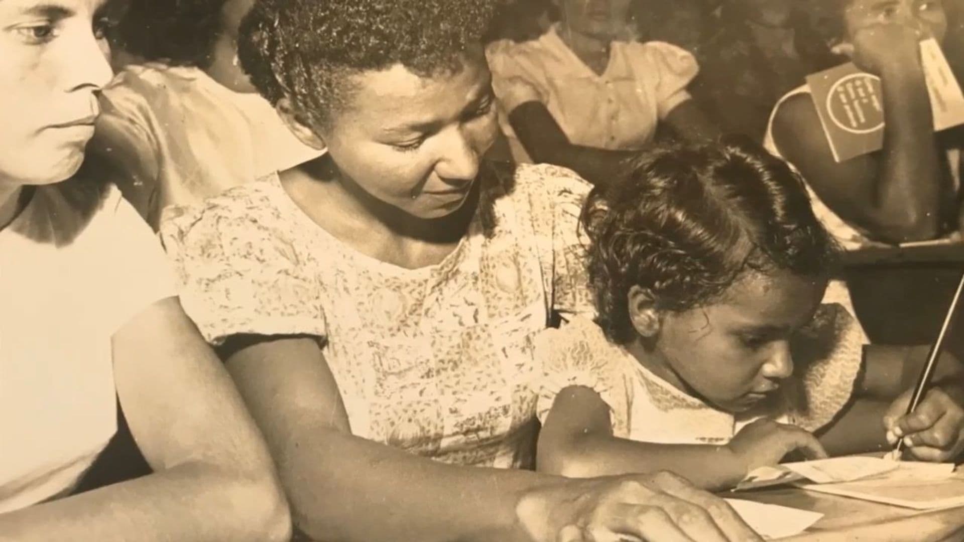 A sepia-toned film still from Reading the World shows a young Brazilian girl sitting on the lap of an older Brazilian woman as they look at a piece of paper on a desk. The girl is holding a pencil and writing on the paper. Around them are other women sitting at desks.