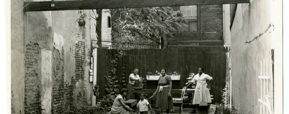 A black-and-white film still from Tenants of Lenapehocking in the Age of Magnets shows a group of four Black women and one Black boy on a lawn. The lawn is surrounded by taller townhomes and blocked from the road with a white picket fence. The group stands around a brick circle with taller foliage.