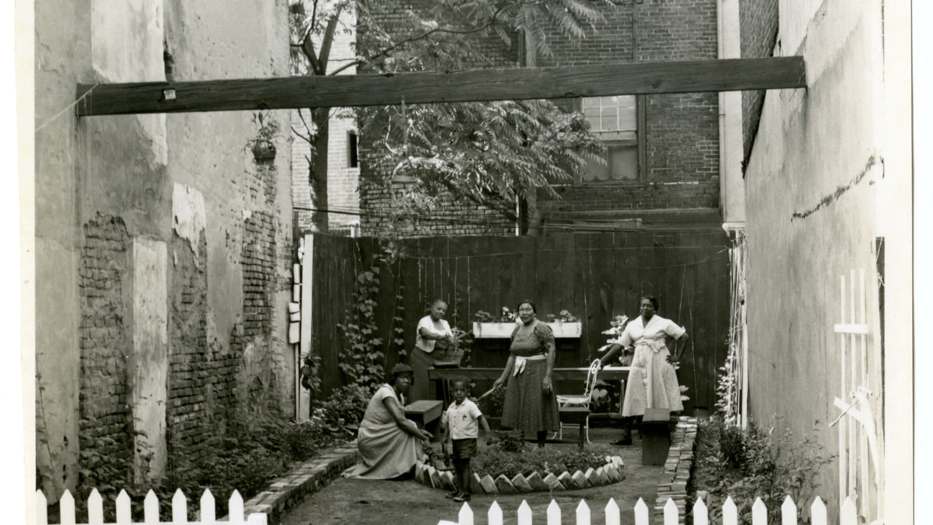 A black-and-white film still from Tenants of Lenapehocking in the Age of Magnets shows a group of four Black women and one Black boy on a lawn. The lawn is surrounded by taller townhomes and blocked from the road with a white picket fence. The group stands around a brick circle with taller foliage.
