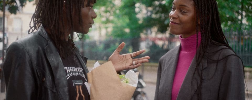A film still from The Aura shows two Black women speaking to each other outside of a park in Paris. The woman on the left is dressed in black and holds a bouquet while the woman on the right wears a pink sweater under a grey blazer.