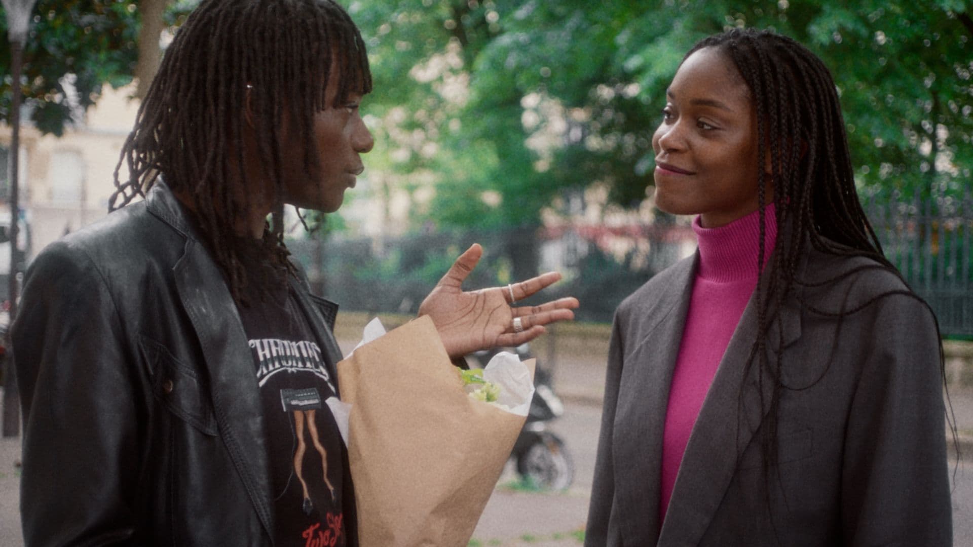 A film still from The Aura shows two Black women speaking to each other outside of a park in Paris. The woman on the left is dressed in black and holds a bouquet while the woman on the right wears a pink sweater under a grey blazer.