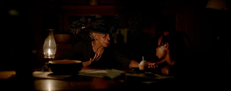 A film still from The Keeper shows an older Black woman speaking to a young Black girl. The two sit around a table, lit by a single gas lantern. The older woman is dressed in all black while the younger girl has a red flower in her hair. On the table in front of them lies a large wooden bowl, some papers, and a cupcake with white frosting.