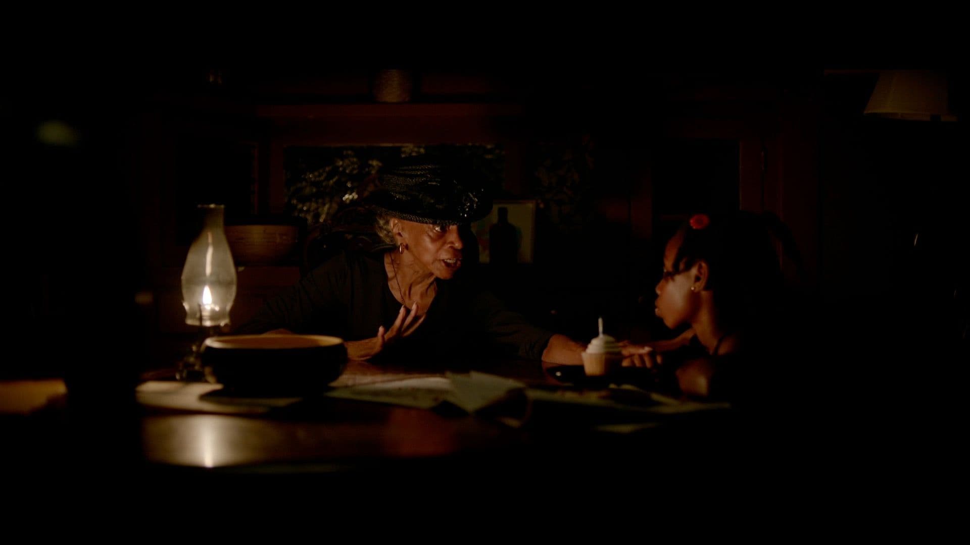 A film still from The Keeper shows an older Black woman speaking to a young Black girl. The two sit around a table, lit by a single gas lantern. The older woman is dressed in all black while the younger girl has a red flower in her hair. On the table in front of them lies a large wooden bowl, some papers, and a cupcake with white frosting.
