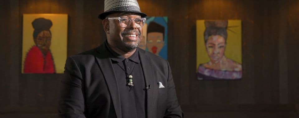 A film still from The Philadelphia Sensei: Lovett Hines shows jazz bassist Christian McBride smiling in front of a wood wall with three portraits of Black women. McBride is a Black man wearing a black shirt and suit jacket, clear glasses, and a black-and white hat.