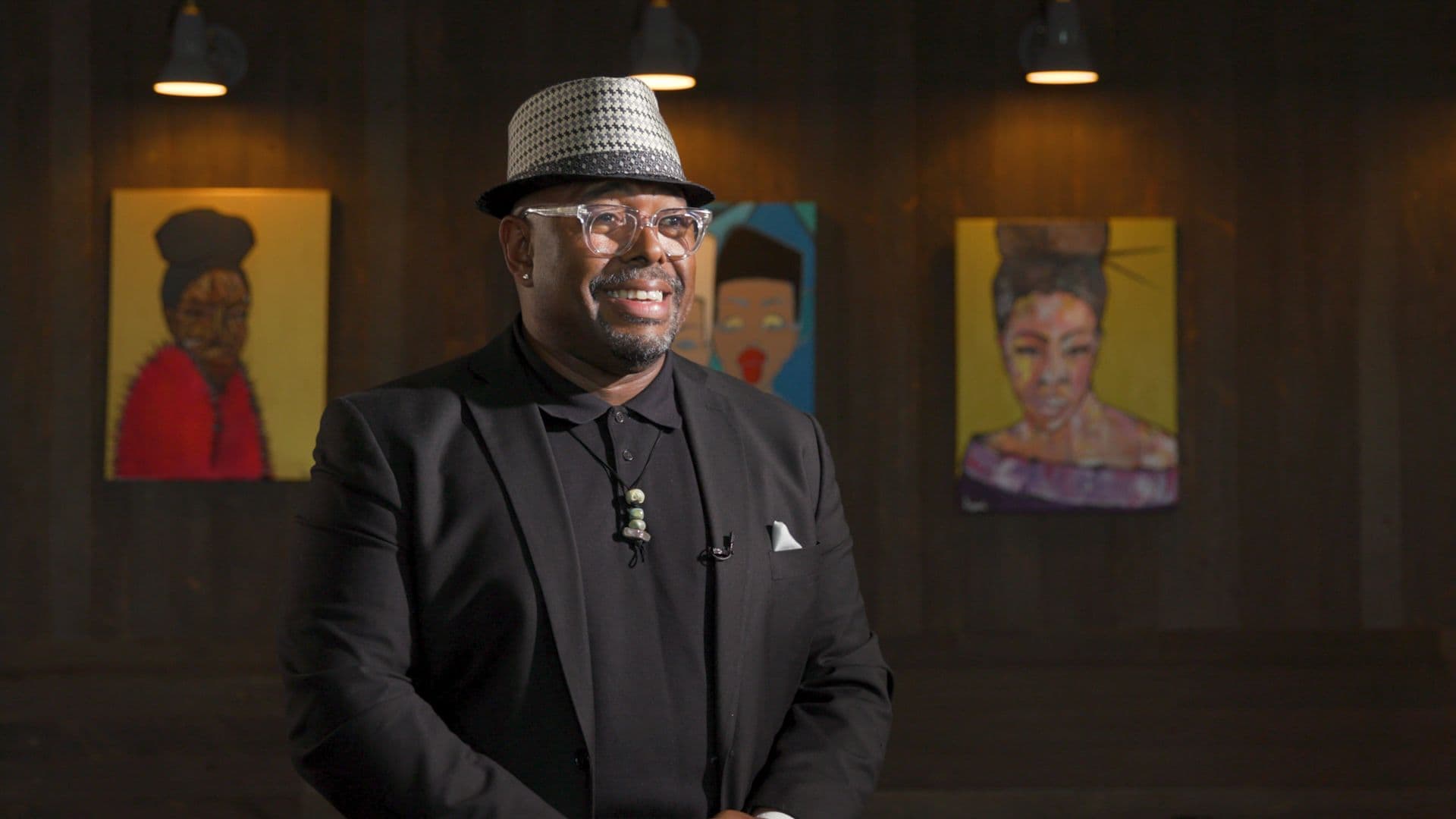 A film still from The Philadelphia Sensei: Lovett Hines shows jazz bassist Christian McBride smiling in front of a wood wall with three portraits of Black women. McBride is a Black man wearing a black shirt and suit jacket, clear glasses, and a black-and white hat.