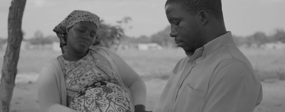 A black-and-white film still from The Prophet shows a pregnant Black woman sitting behind a Black man. The woman is facing the front and looks down solemnly. She wears a patterned dress and bandana. The man is facing the left and also looks down. He is wearing a light-toned dress shirt.