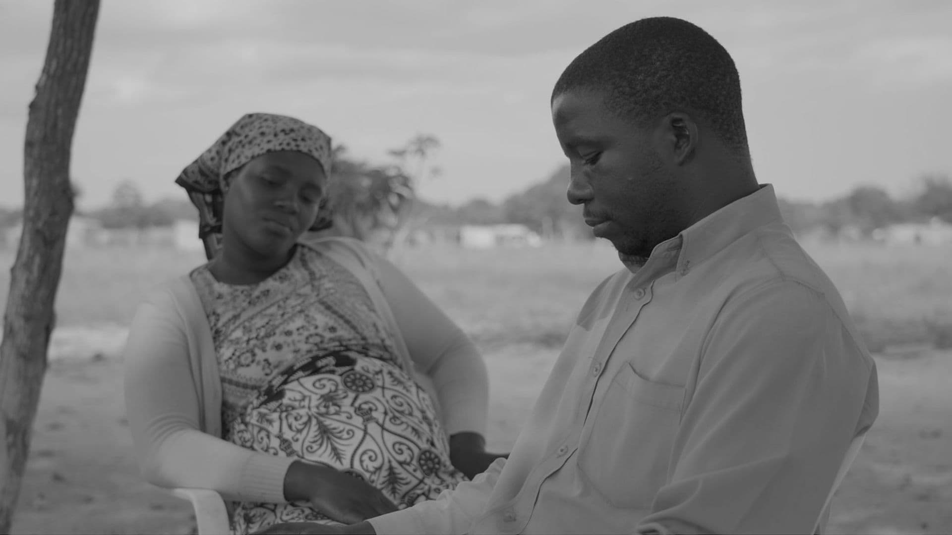 A black-and-white film still from The Prophet shows a pregnant Black woman sitting behind a Black man. The woman is facing the front and looks down solemnly. She wears a patterned dress and bandana. The man is facing the left and also looks down. He is wearing a light-toned dress shirt.