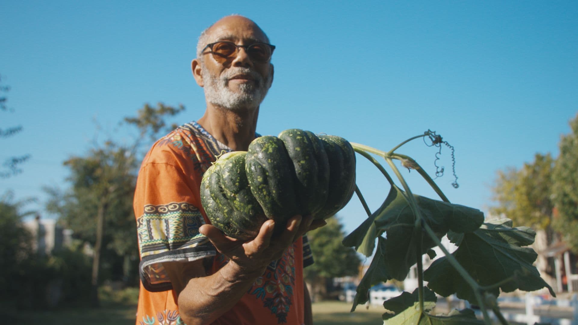 A film still from The Seeds We Carry shows an older Haitian man holding a green, speckled pumpkin in his right hand. The man wears a patterned orange shirt and tinted sunglasses. He stands in front of a large patch of grass blocked off from the street with a white fence.