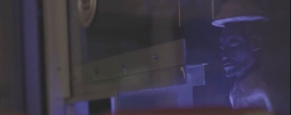 A film still from The Store shows a small stone statue behind a clear barrier. The barrier has a dark sign nailed into it and a small opening.