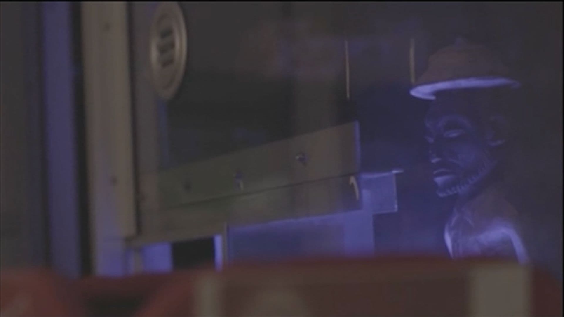 A film still from The Store shows a small stone statue behind a clear barrier. The barrier has a dark sign nailed into it and a small opening.