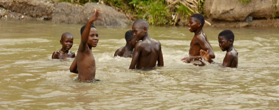 A film still from The Tropic Sun and His Eyes shows a group of six shirtless Black boys playing in a body of water. One boy raises his right arm as he looks off into the distance while the rest of the group look around at each other.
