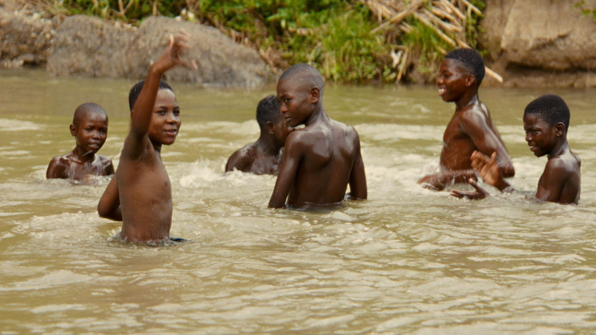 A film still from The Tropic Sun and His Eyes shows a group of six shirtless Black boys playing in a body of water. One boy raises his right arm as he looks off into the distance while the rest of the group look around at each other.