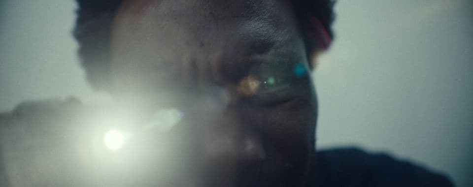A film still from To Run is to Return shows a Black man leaning down, holding a flashlight directly towards the camera. The light gives off a lens flare that blocks the man’s eyes.