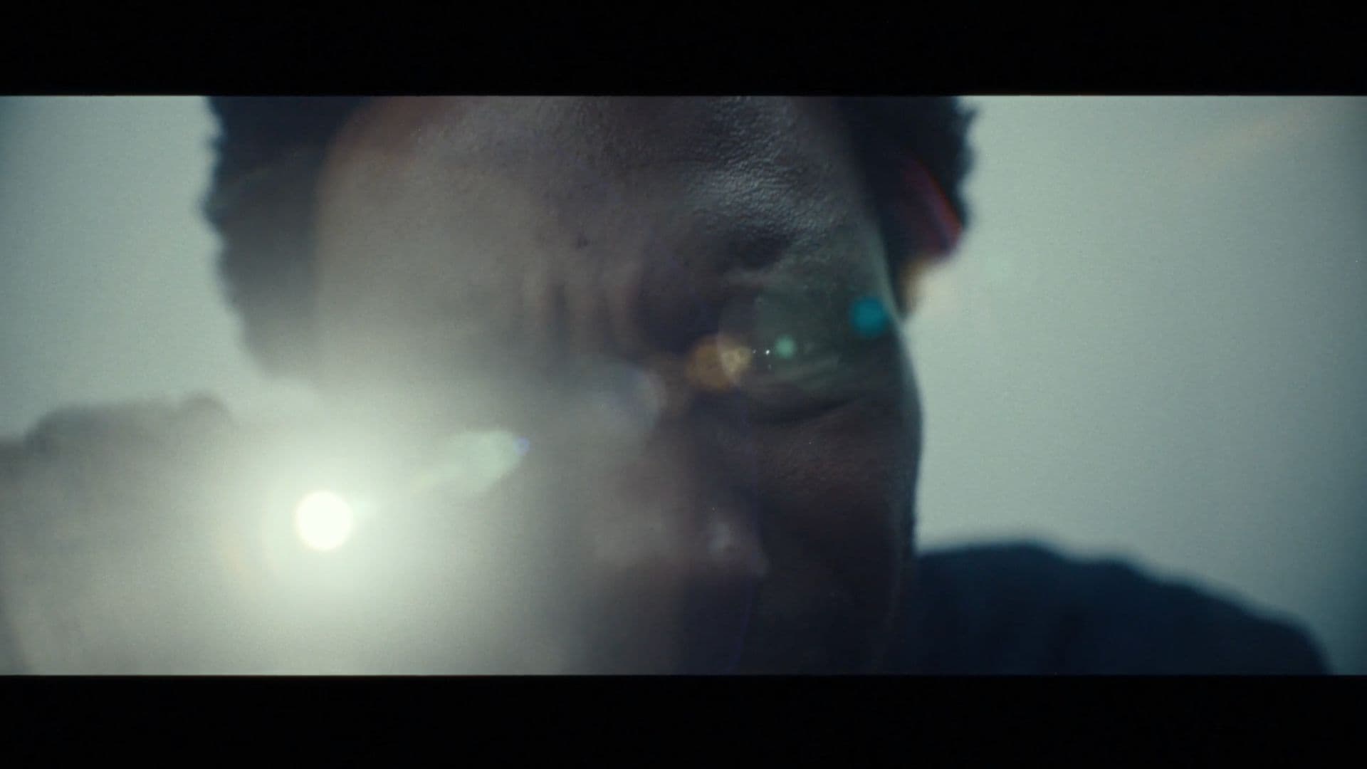 A film still from To Run is to Return shows a Black man leaning down, holding a flashlight directly towards the camera. The light gives off a lens flare that blocks the man’s eyes.