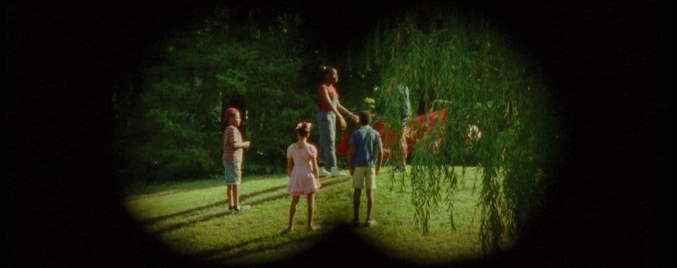 A film still from Treasure Island shows two boys and two girls in a park through a binocular overlay. The boy on the left wears a red cap, a red-and-white striped shirt, and jean shorts. The girl in the middle wears a pink dress with bows in her hair. The boy on the right wears a blue shirt and khaki shorts. They all stand and look towards the taller girl in a red shirt and blue overalls, who is facing to the right and holding a large piece of red fabric.