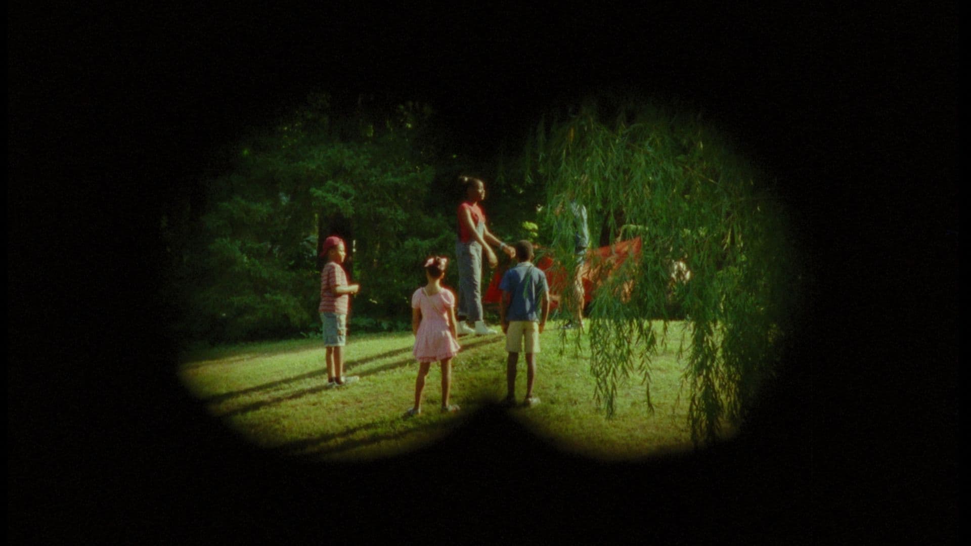 A film still from Treasure Island shows two boys and two girls in a park through a binocular overlay. The boy on the left wears a red cap, a red-and-white striped shirt, and jean shorts. The girl in the middle wears a pink dress with bows in her hair. The boy on the right wears a blue shirt and khaki shorts. They all stand and look towards the taller girl in a red shirt and blue overalls, who is facing to the right and holding a large piece of red fabric.