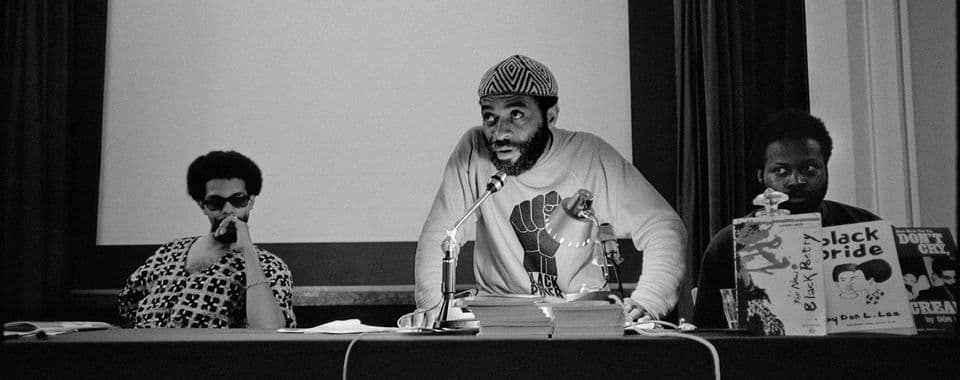 A black-and-white film still from True North shows a Black man in a patterned hat and a “Black Power” shirt leaning over a microphone. The man sits between two other Black men on a panel. Next to him are books that read, “The New Black Poetry,” “Black Pride,” and “Don’t Cry, Scream.”