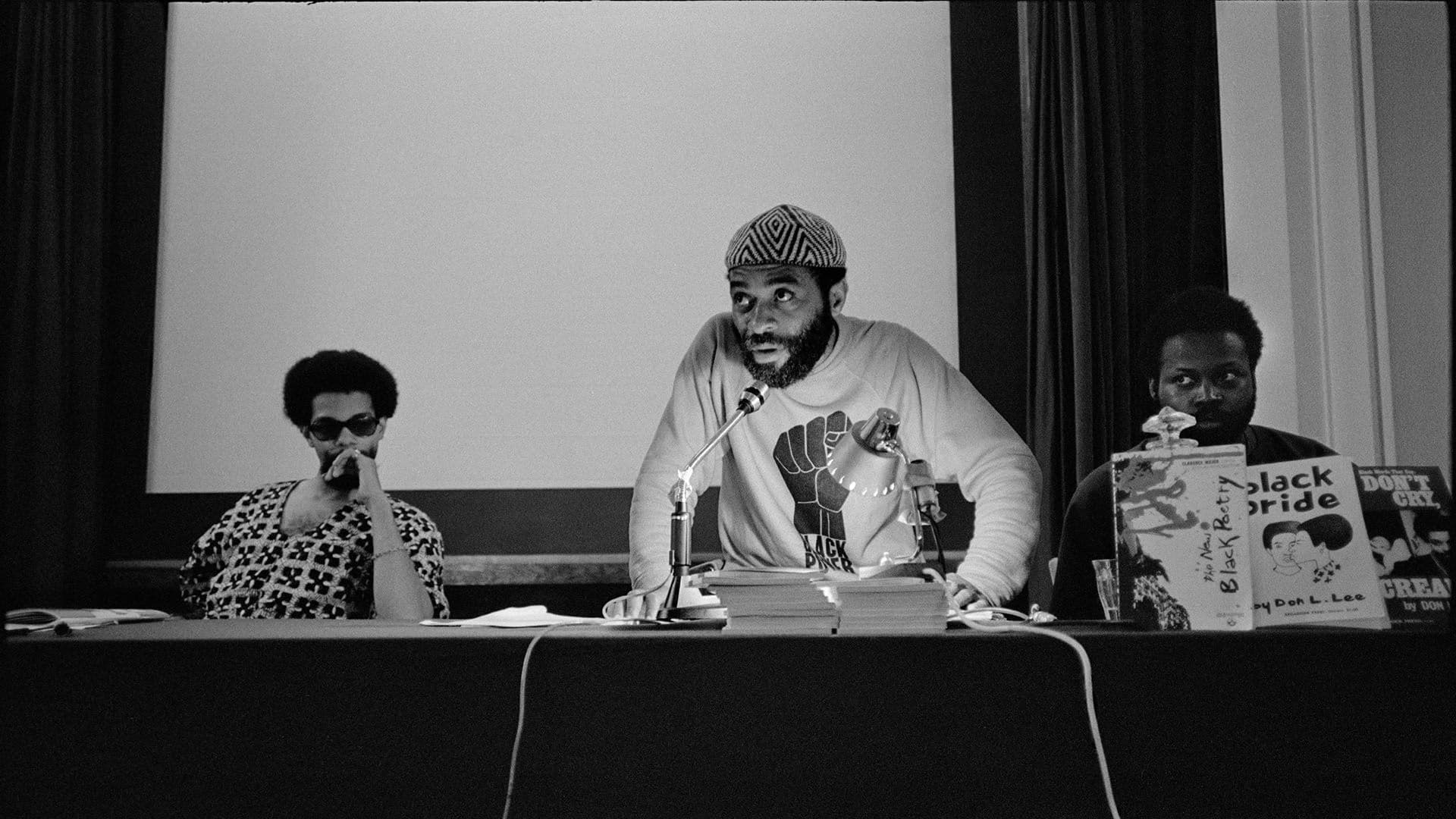 A black-and-white film still from True North shows a Black man in a patterned hat and a “Black Power” shirt leaning over a microphone. The man sits between two other Black men on a panel. Next to him are books that read, “The New Black Poetry,” “Black Pride,” and “Don’t Cry, Scream.”