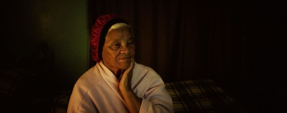 A film still from Variations on a Theme shows an older Black woman in a pale robe and red bonnet staring pensively at a television. Her left hand rests on her chin as she sits in a dimly lit bedroom.