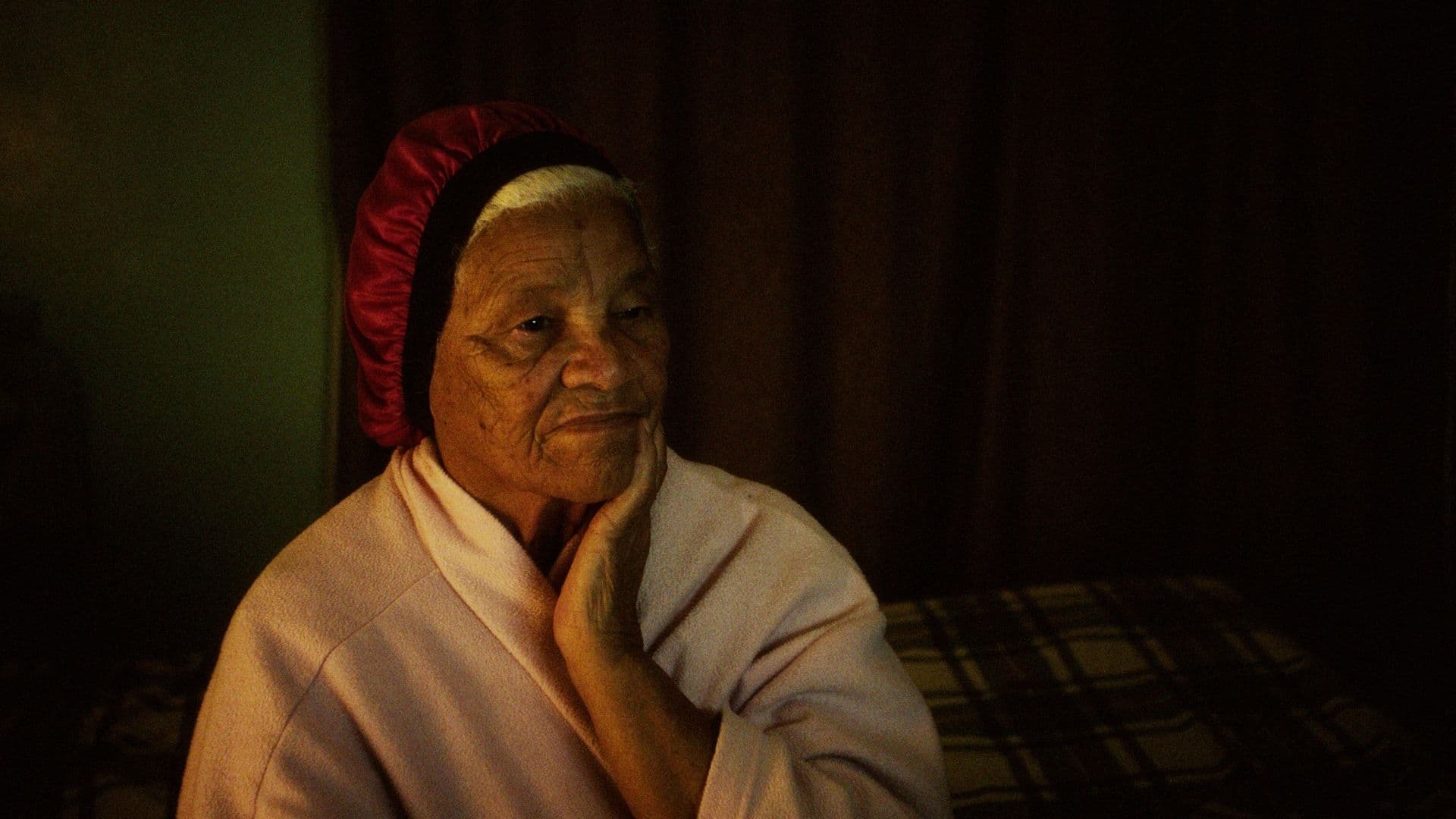 A film still from Variations on a Theme shows an older Black woman in a pale robe and red bonnet staring pensively at a television. Her left hand rests on her chin as she sits in a dimly lit bedroom.