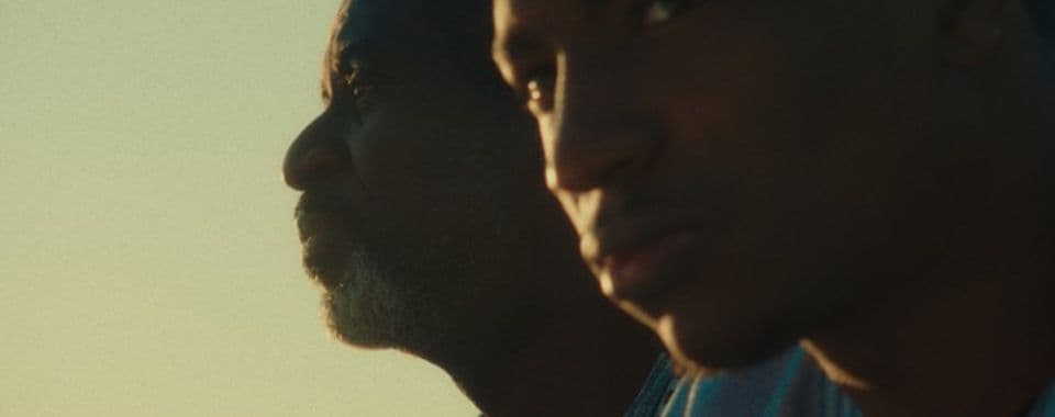 A film still from Vis-à-Vis shows two Black men facing the left with the sun reflecting off their face. The younger man is closer to the camera and stares off to the side, while the older man behind him stares straight left.