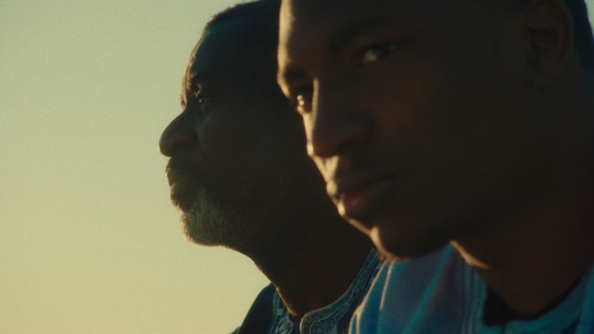 A film still from Vis-à-Vis shows two Black men facing the left with the sun reflecting off their face. The younger man is closer to the camera and stares off to the side, while the older man behind him stares straight left.