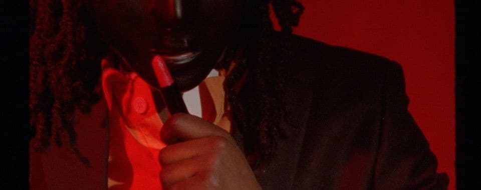 A film still from Voicemail shows a close-up of a Black person under red lighting wearing a dark mask and a jacket with a collared shirt and tie. They are holding up a tube of red lipstick to their face.