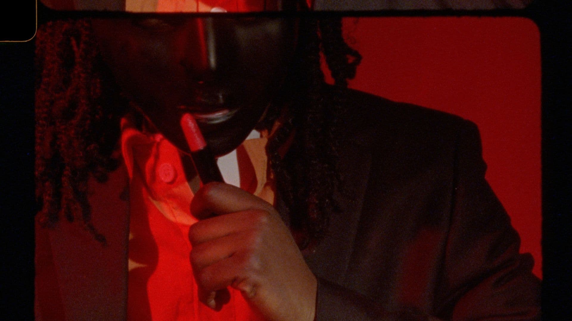 A film still from Voicemail shows a close-up of a Black person under red lighting wearing a dark mask and a jacket with a collared shirt and tie. They are holding up a tube of red lipstick to their face.