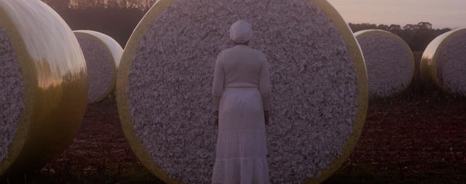 A film still from Watch Out for the Ghosts shows a Black woman in a white dress standing in front of a cotton bale. She is facing away from the camera. There are more cotton bales around her and some trees in the distance.