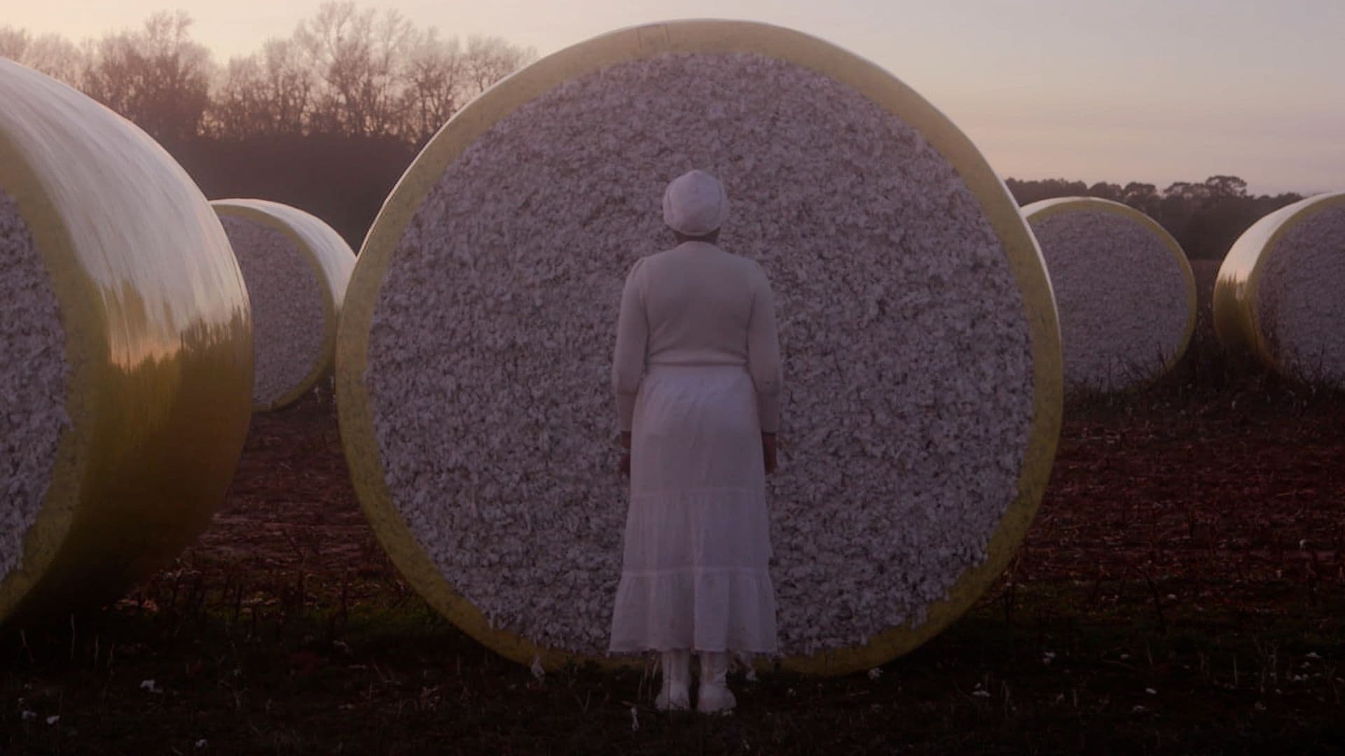 A film still from Watch Out for the Ghosts shows a Black woman in a white dress standing in front of a cotton bale. She is facing away from the camera. There are more cotton bales around her and some trees in the distance.