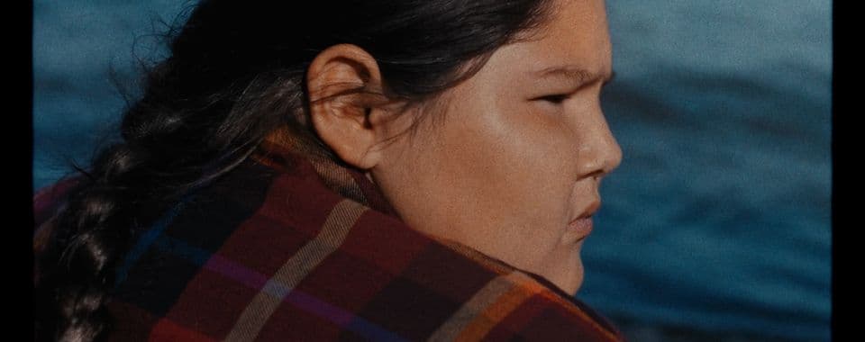 A film still from We Act Like Children shows two Anishinaabe children sitting in a brown tent. The girl on the right wears a red plaid flannel shirt and has her hair pulled back in a braid. She is looking towards the older boy on the left who is facing the camera. He wears a teal plaid flannel over a green shirt. On the floor of the tent, there’s a small table and a collection of colorful blankets.