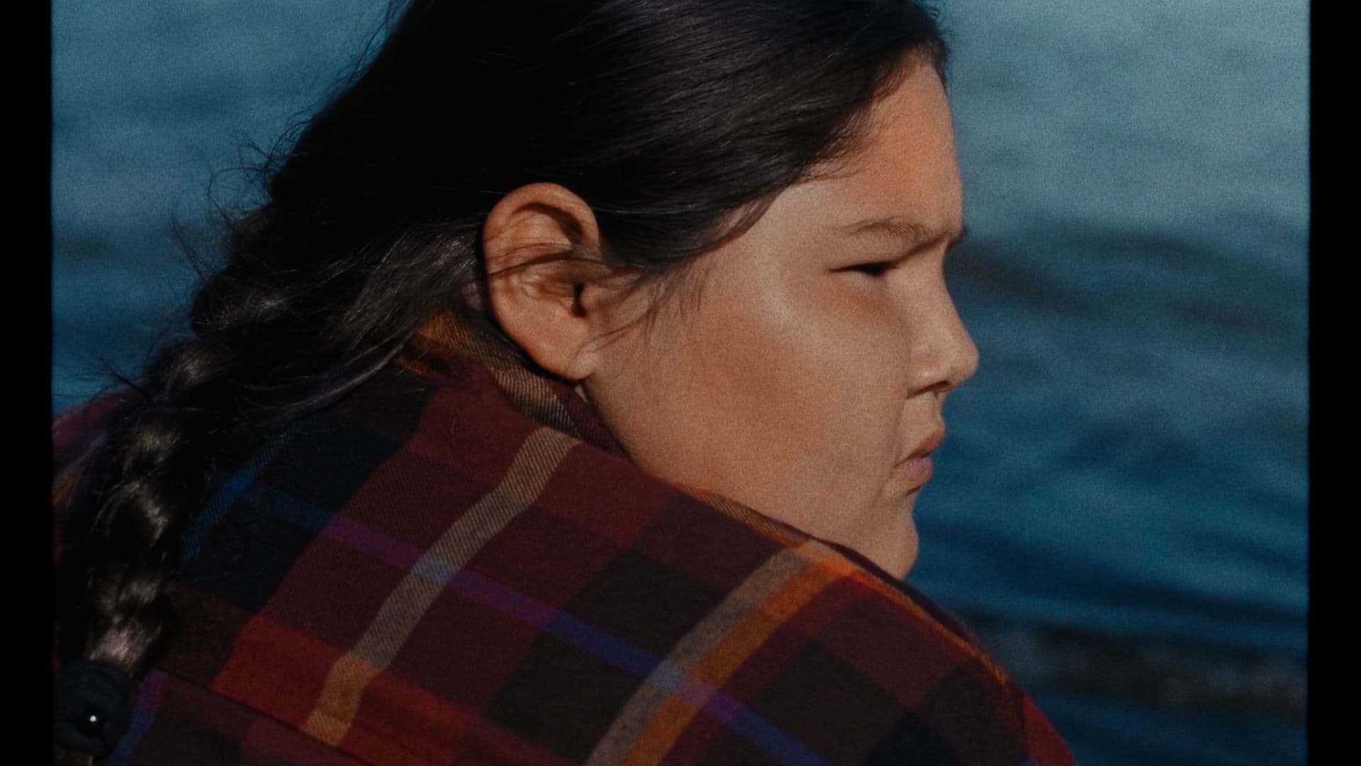 A film still from We Act Like Children shows two Anishinaabe children sitting in a brown tent. The girl on the right wears a red plaid flannel shirt and has her hair pulled back in a braid. She is looking towards the older boy on the left who is facing the camera. He wears a teal plaid flannel over a green shirt. On the floor of the tent, there’s a small table and a collection of colorful blankets.
