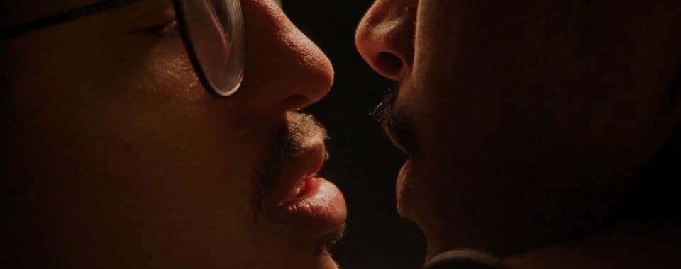A film still from We Are Not Dreaming shows a close-up of two young Black men with their faces close, about to kiss. The man on the left wears glasses while the man on the right has his arm embracing the other man.