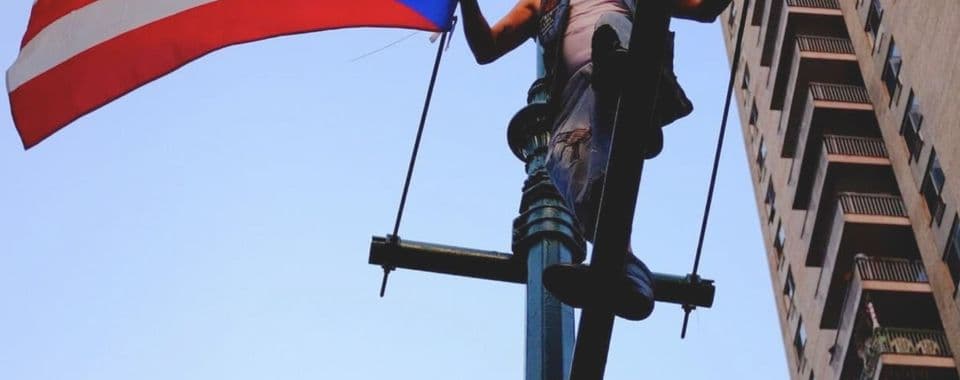A film still from West Side Familia shows an older Puerto Rican man stands on top of a traffic light pole in New York City, hanging a Puerto Rican flag. The man is wearing a white tank top under a leather vest. Behind him, a tall apartment building stretches into the sky.