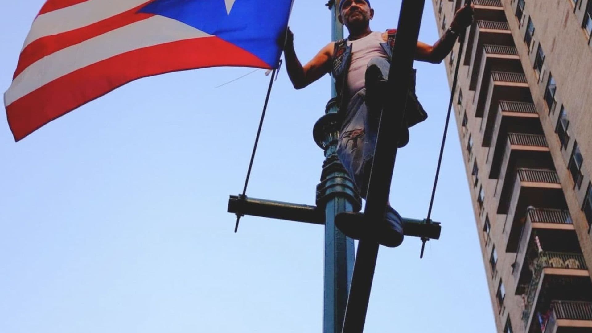 A film still from West Side Familia shows an older Puerto Rican man stands on top of a traffic light pole in New York City, hanging a Puerto Rican flag. The man is wearing a white tank top under a leather vest. Behind him, a tall apartment building stretches into the sky.