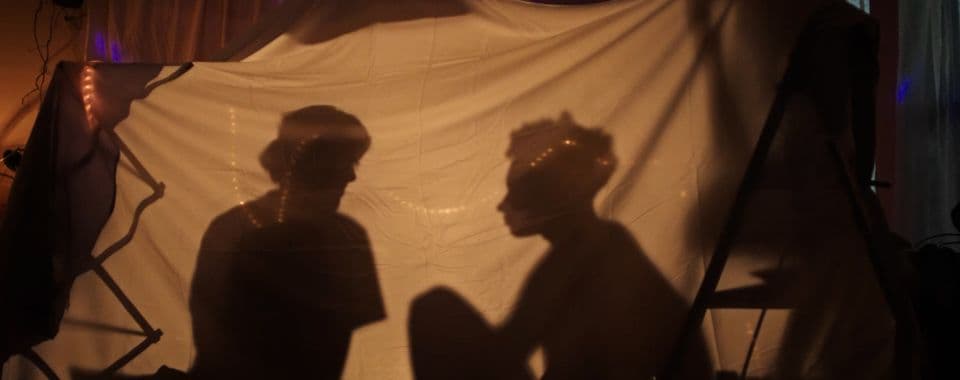 A film still from What Will I Become? shows two silhouettes inside a tent made from bedsheets. The tent is lit by strings of warm lights.
