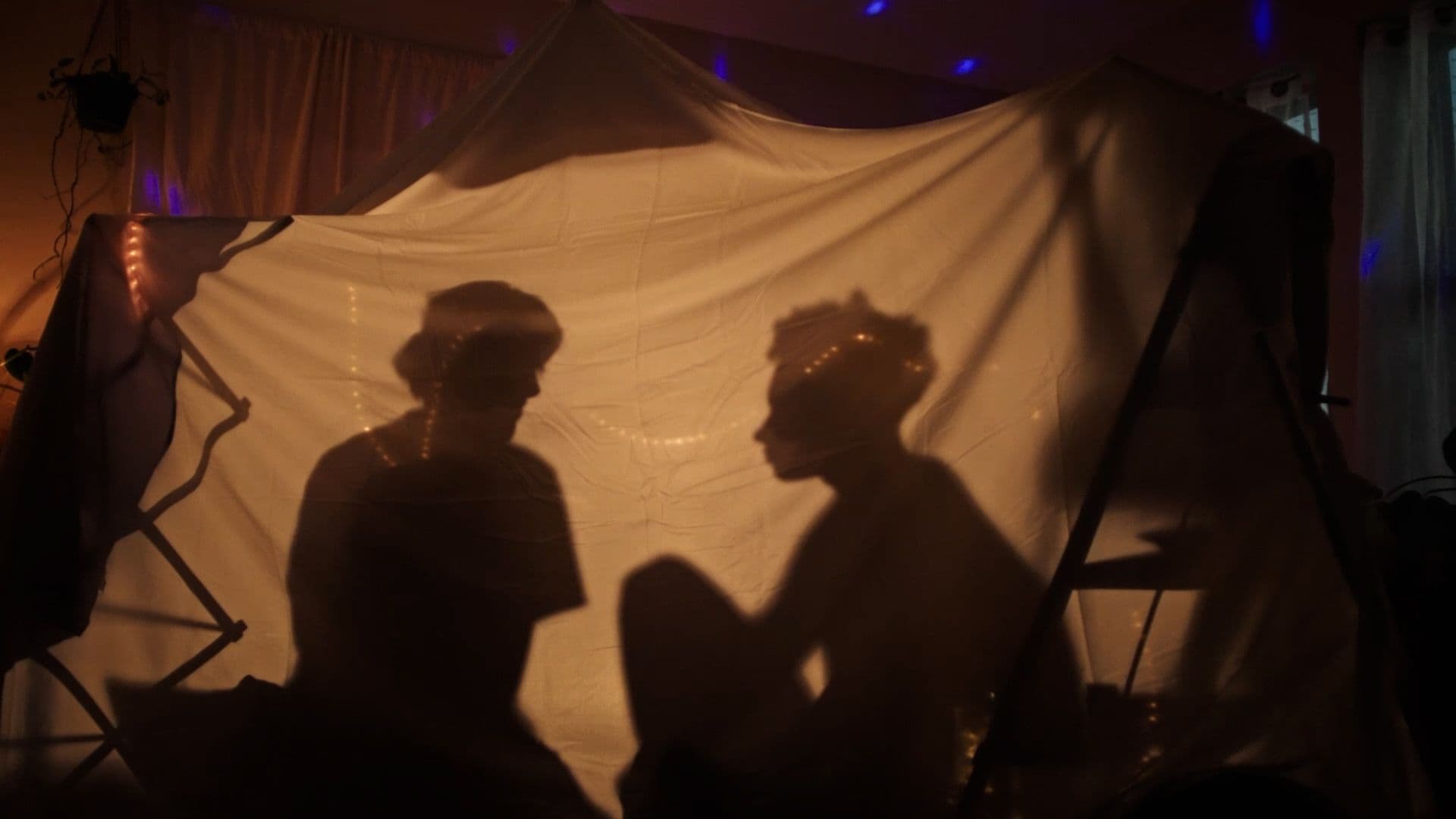 A film still from What Will I Become? shows two silhouettes inside a tent made from bedsheets. The tent is lit by strings of warm lights.