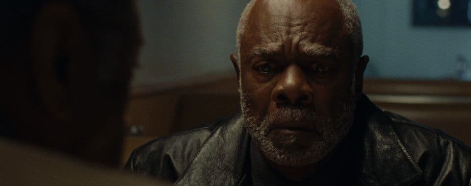 A film still from When They’re Gone shows an older Black man in a black leather jacket sitting in a booth across from another Black man. The older man has his fingers interlocked and is looking down in distress.