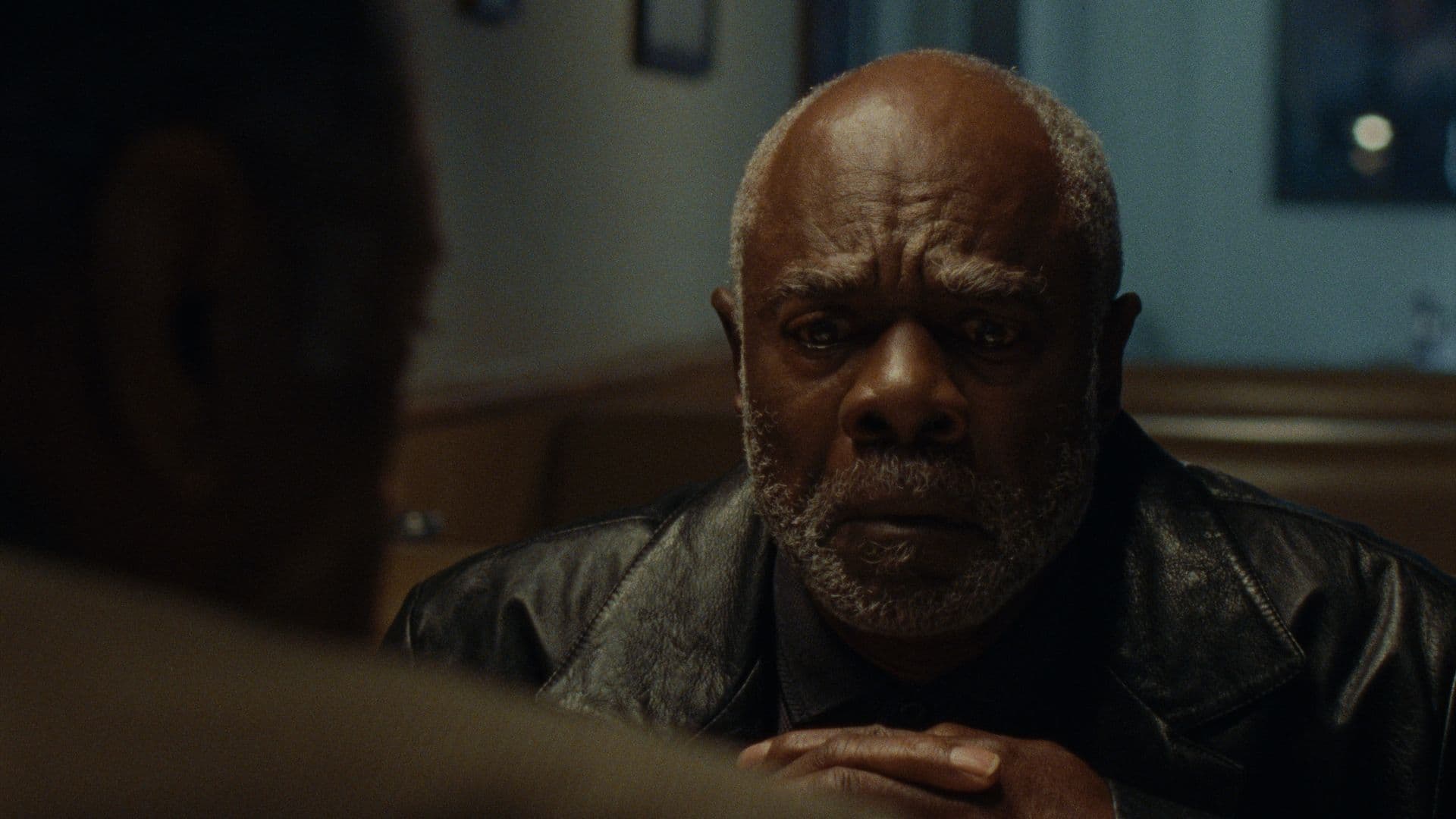 A film still from When They’re Gone shows an older Black man in a black leather jacket sitting in a booth across from another Black man. The older man has his fingers interlocked and is looking down in distress.