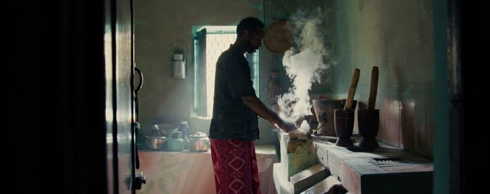 A film still from Whispers of a Burning Scent shows a Black man in a blue shirt and patterned red bottoms standing in a kitchen. He is facing right and holds a smoking dish. Around him are various pots and kettles, as well as many large stone mortars and pestles.