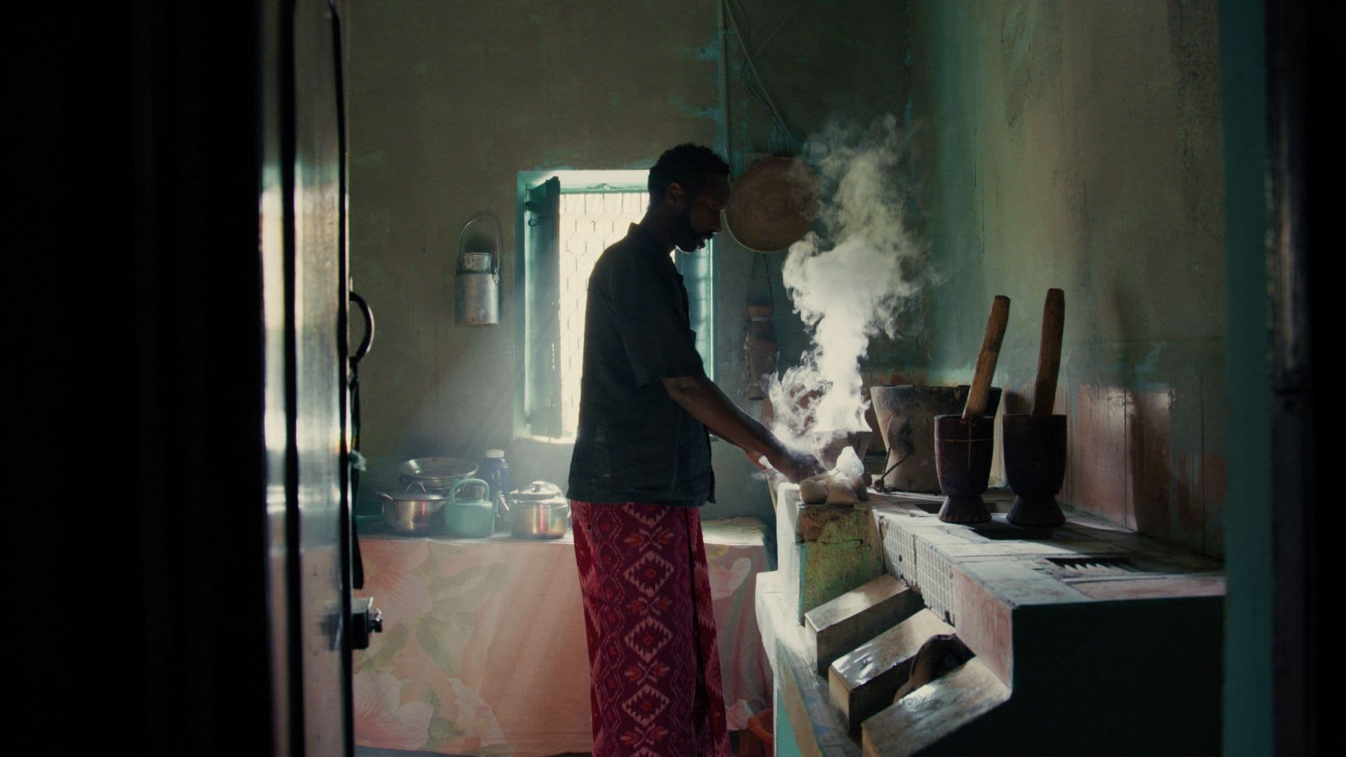 A film still from Whispers of a Burning Scent shows a Black man in a blue shirt and patterned red bottoms standing in a kitchen. He is facing right and holds a smoking dish. Around him are various pots and kettles, as well as many large stone mortars and pestles.