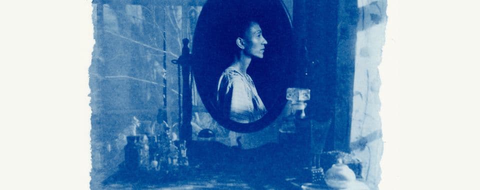 A blue-toned film still from White Musk shows a cluttered vanity with an oval-shaped mirror. In the mirror, there’s a reflection of a woman facing right.