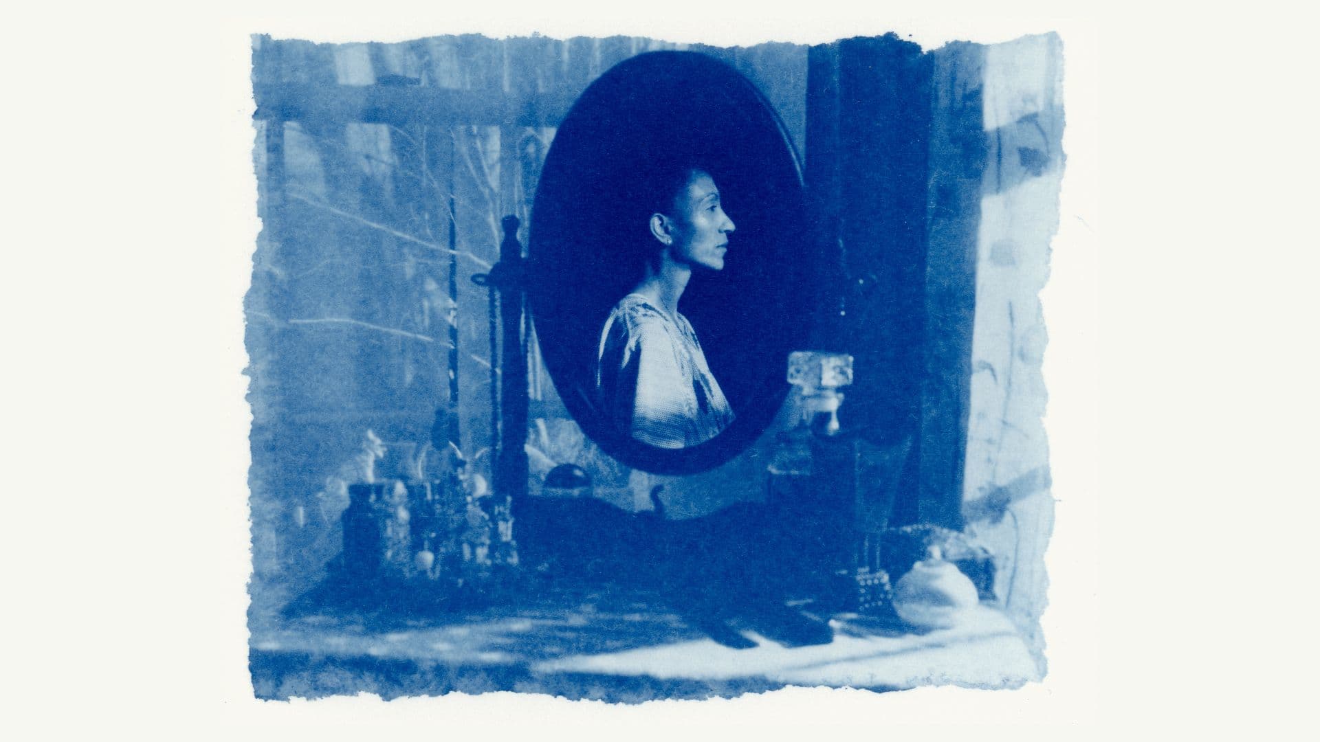 A blue-toned film still from White Musk shows a cluttered vanity with an oval-shaped mirror. In the mirror, there’s a reflection of a woman facing right.