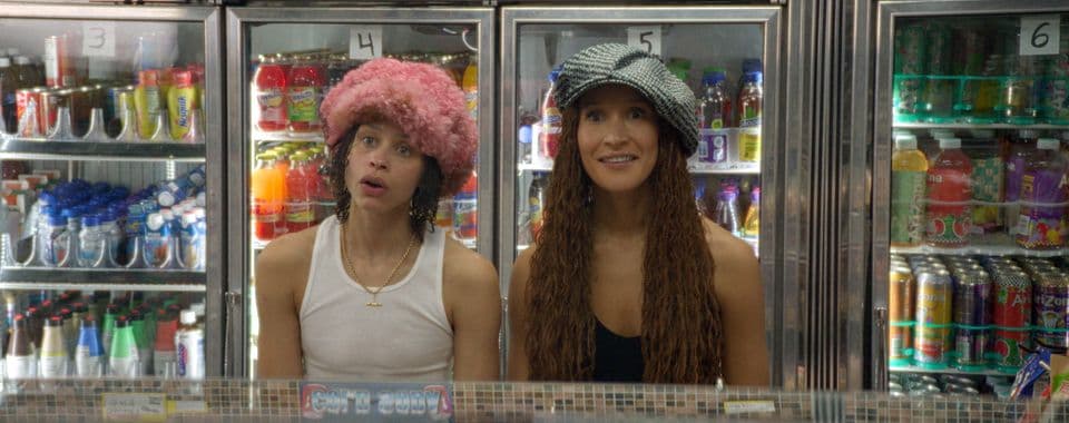 A film still from Wholesome shows two Black women standing in front of a deli counter. The woman on the left wears a fluffy pink hat, a gold chain necklace, and a white tank top. The woman on the right wears an oversized black-and-white beret and a black tank top. They both look forward in awe. Behind them, there’s four refrigerators displaying various types of beverages.