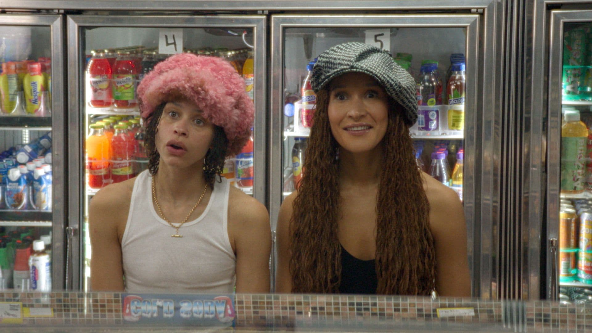 A film still from Wholesome shows two Black women standing in front of a deli counter. The woman on the left wears a fluffy pink hat, a gold chain necklace, and a white tank top. The woman on the right wears an oversized black-and-white beret and a black tank top. They both look forward in awe. Behind them, there’s four refrigerators displaying various types of beverages.