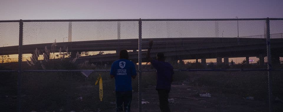 A film still from Wood Street shows two Black men standing in front of a chain-link fence, looking at an empty lot and a distant highway ramp. The man on the left wears a blue shirt and the man on the right has his left arm raised, holding onto the fence. The sky above them is a dark orange and purple.
