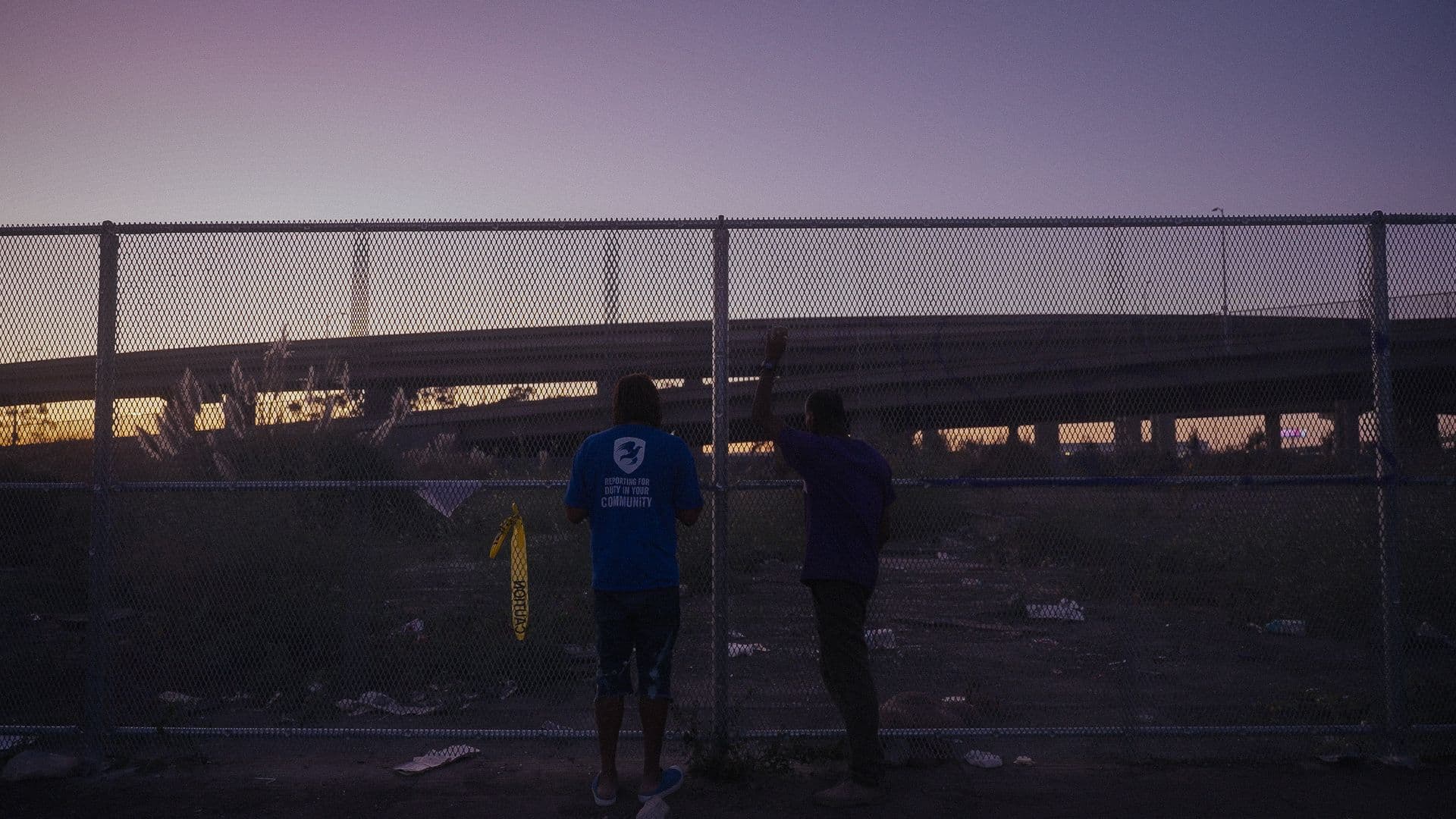 A film still from Wood Street shows two Black men standing in front of a chain-link fence, looking at an empty lot and a distant highway ramp. The man on the left wears a blue shirt and the man on the right has his left arm raised, holding onto the fence. The sky above them is a dark orange and purple.