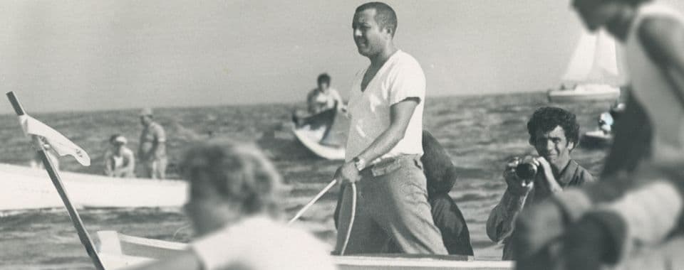 A black-and-white film still from Zenón and the Rebel Boats shows Carlos Zenón standing on a small boat in the Caribbean sea. Zenón is a Puerto Rican man in a white shirt. Around him, there are many other fishermen in boats.