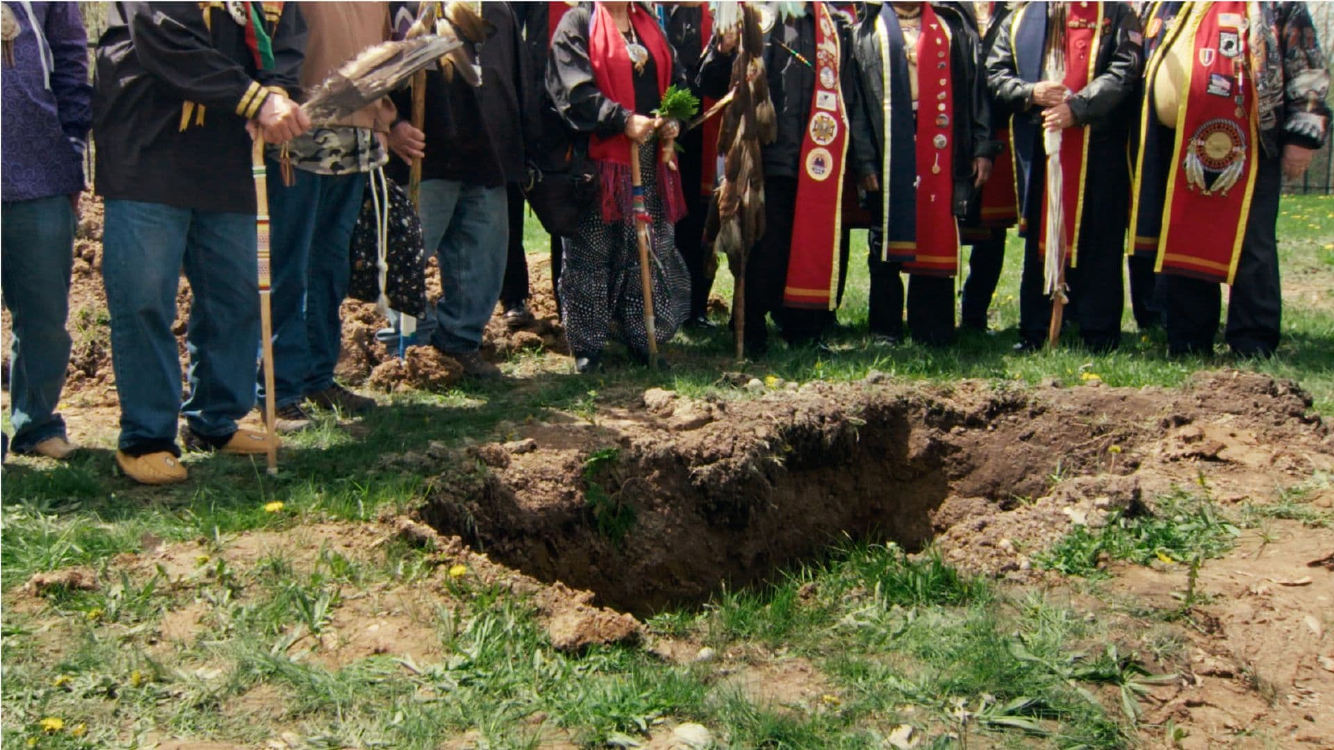 A film still from Aanikoobijigan (ancestor/great-grandparent/great-grandchild) shows a group of Native people standing around a freshly dug rectangular pit on a sunny day. The frame is centered on the ground, showing the grass, the pit, and the lower bodies of the people.