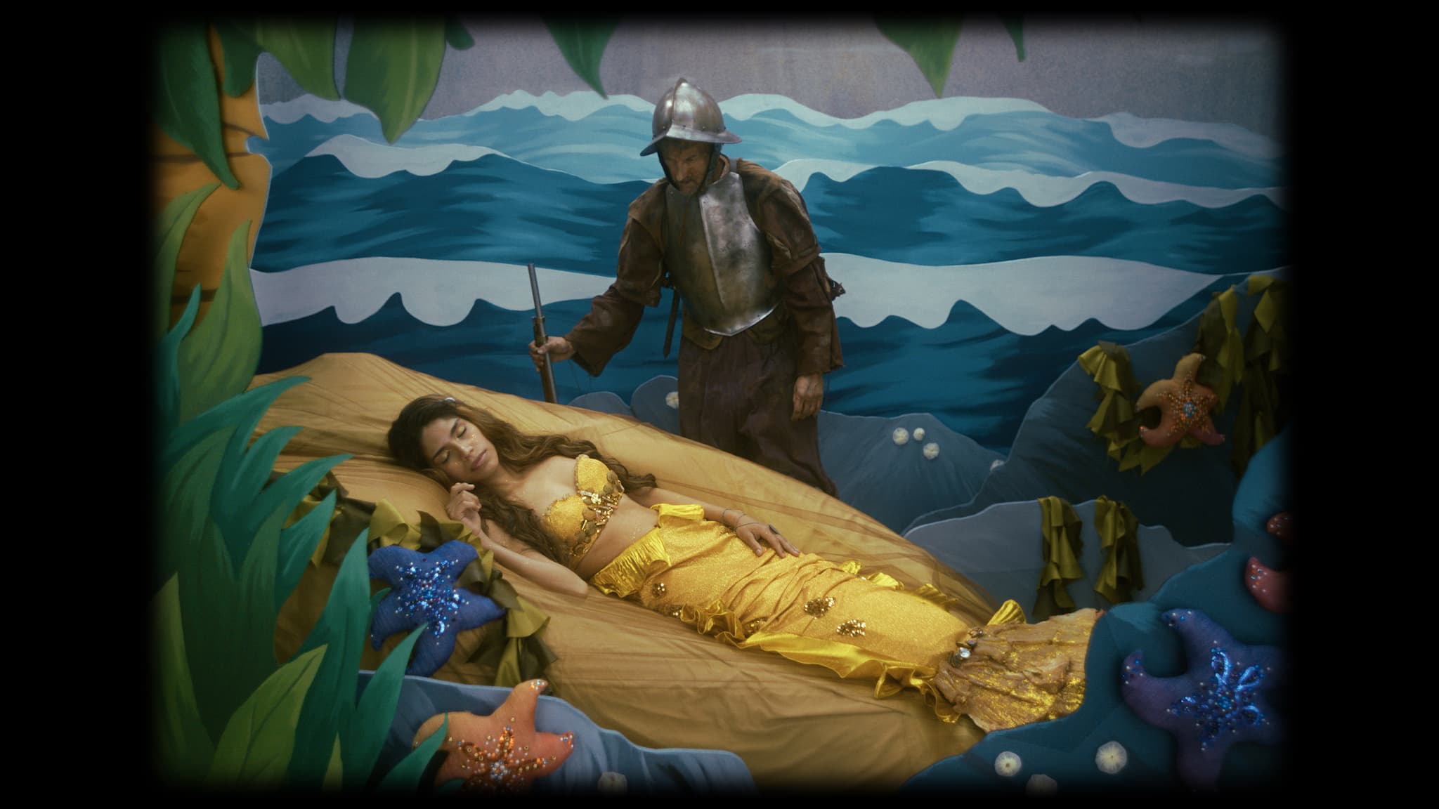 A film still from Las Cosas Que Brillan shows a black frame around a staged scene. Inside the frame is a woman dressed as a mermaid, with a yellow tail and top, sleeping on a faux rock. Standing above her is a man dressed as a soldier, wearing dirty armor with a grimy face, gazing down at her. They are positioned within a theatrical set designed to mimic an ocean scene with a series of waves behind them.